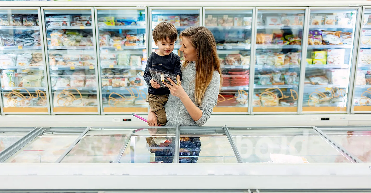 A mother is carrying her son and both are holding a frozen packaged item in the frozen section of a grocery store.